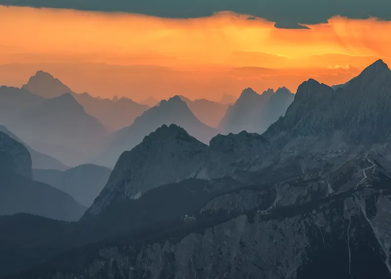 View over a layered mountain range at sunset, with an orange sky and mountains in blue and grey tones.
