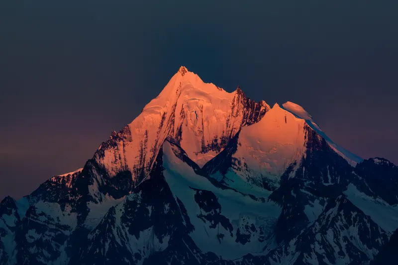 A snow-capped mountain peak glows orange in the sunlight, while the rest of the mountain range and sky are dark blue.
