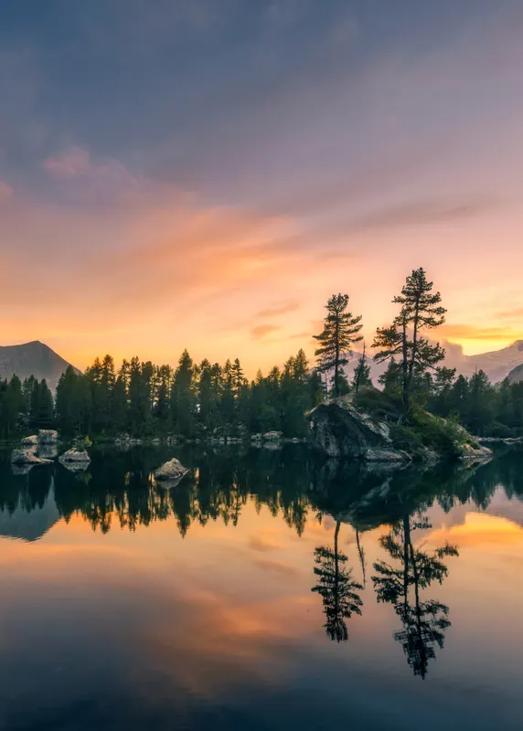 A calm alpine lake at sunset, featuring a small rocky island with pine trees reflected in the water. The sky is orange and blue.