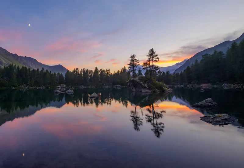 Mountain lake at sunset with pink and orange sky, surrounded by forests and mountains, featuring a small island and moon.