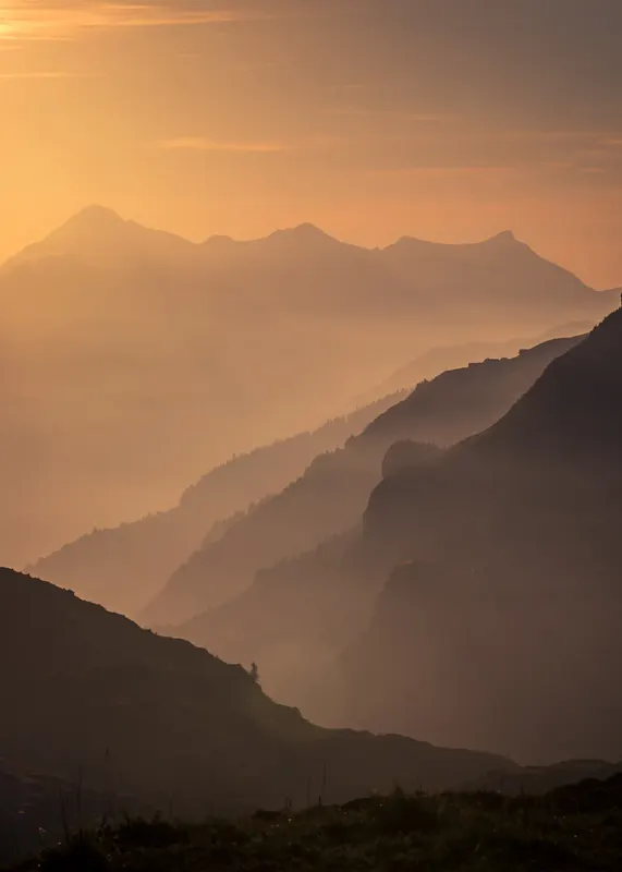 A mountain panorama at sunrise with multiple layers of mountains bathed in warm, golden light and haze.