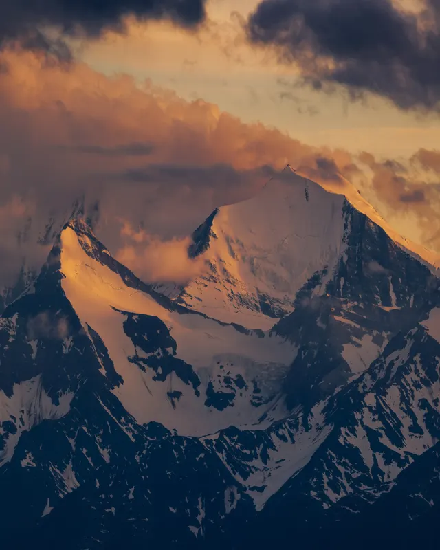 Snow-capped alpine peaks bathed in warm twilight, enveloped by golden and dark clouds.