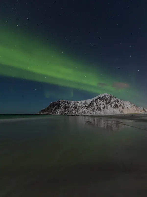 Ein grünes Polarlichtband erstreckt sich über einen sternenklaren Nachthimmel, der über einem schneebedeckten Berg und einem ruhigen Strand liegt.