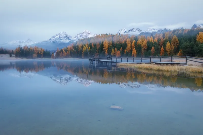 A still mountain lake reflecting snow-capped mountains and golden larch forests. A wooden pier extends into the water, light mist.