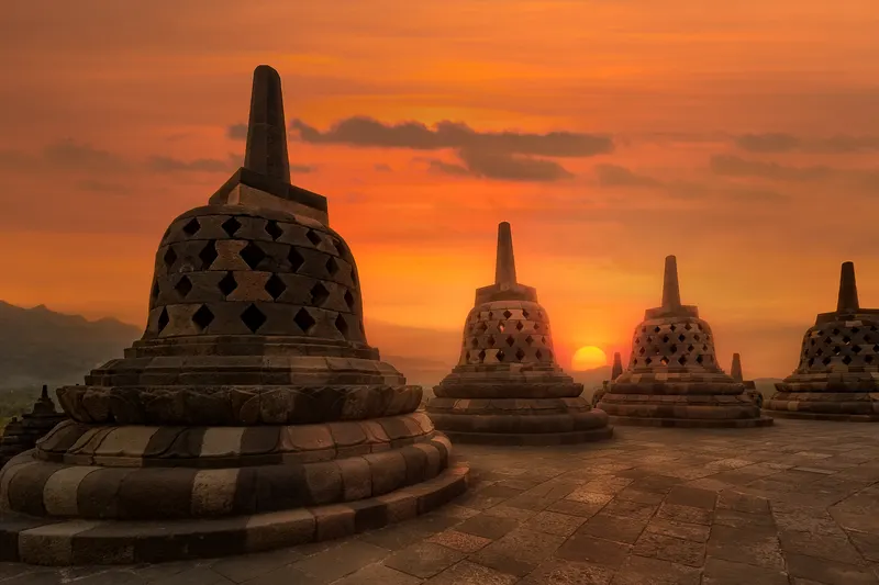 Mehrere alte Stein-Stupas des Borobudur-Tempels bei Sonnenuntergang, mit einem orangefarbenen Himmel und Bergen im Hintergrund.