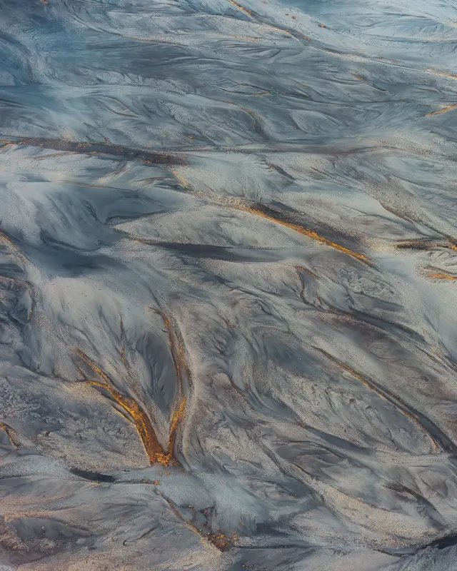 Luftbild eines geflochtenen Flusses in Island mit grauen Sedimenten und goldenen Wasserläufen, die abstrakte Muster bilden.