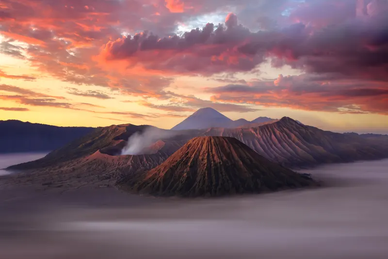 Panoramic view of a volcanic landscape at sunrise, featuring a smoking volcano, multiple volcanic cones, and vibrant red clouds.