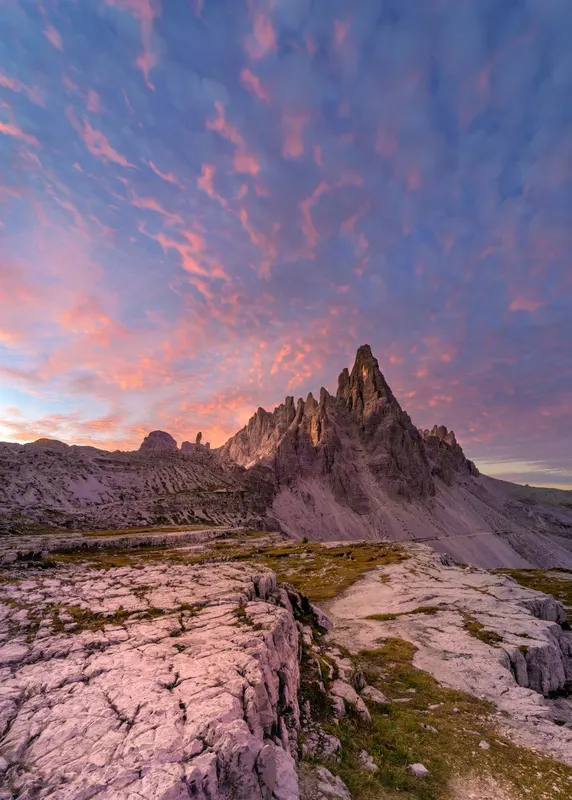A wide landscape of the Dolomites at sunrise featuring rugged mountain peaks and a sky full of pink and blue clouds.