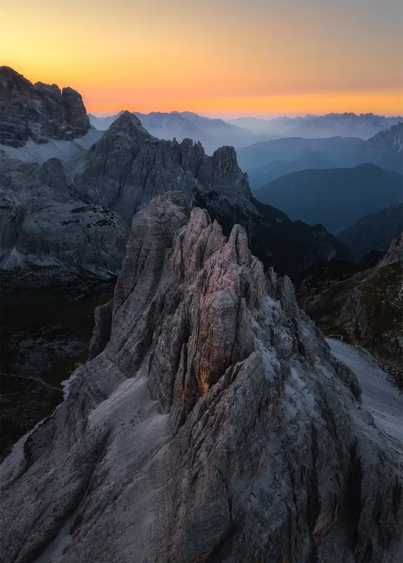 A wide view of rugged Dolomites peaks at sunset, with an orange sky and blue mountain layers in the background.