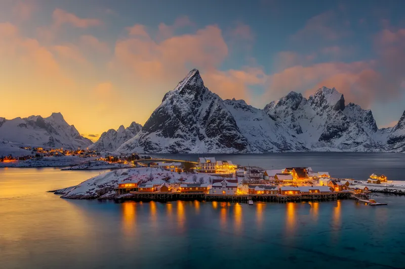 A winter village with illuminated houses by the water, surrounded by snow-covered mountains under an orange and blue twilight sky.