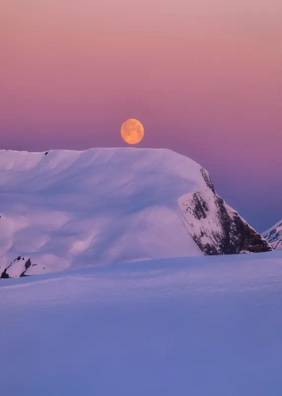 A large, orange full moon rises above a snow-covered mountain at twilight, with a sky in pink and violet hues.