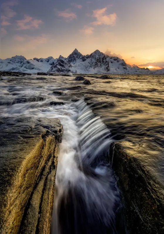 Langzeitbelichtung von Meerwasser, das über Felsen fliesst, mit schneebedeckten Bergen und einem Sonnenuntergangshimmel im Hintergrund.