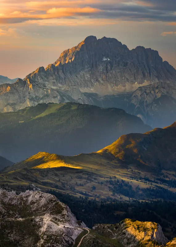 View of the Dolomites at sunset. Rugged mountains, green slopes, and a winding path bathed in warm light.