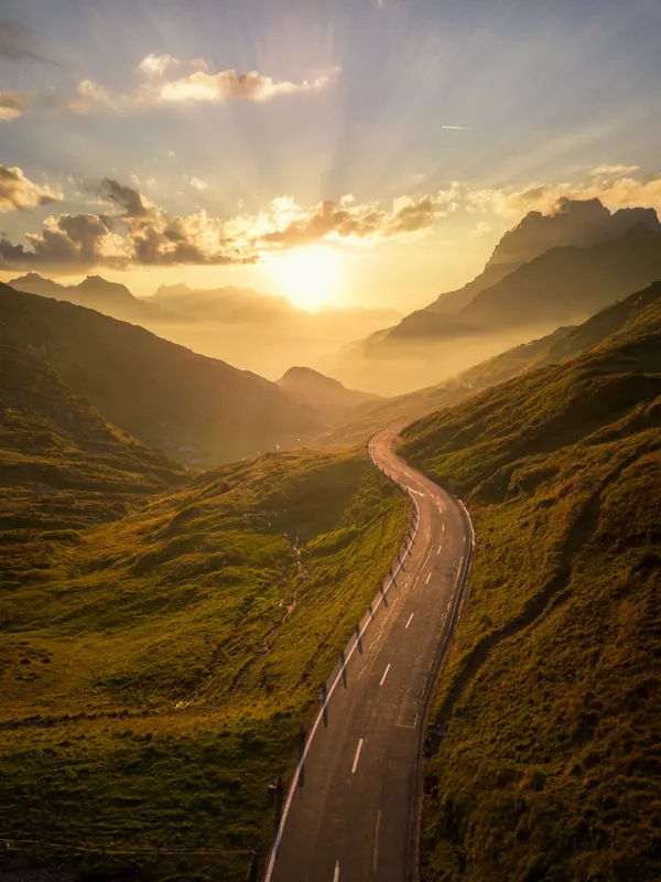 A winding mountain road at sunset. Golden sunbeams break through clouds over a misty valley with distant mountains.