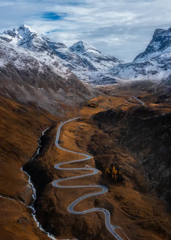 Aerial view of a winding switchback road in the Julier Pass valley, surrounded by autumn foliage and snow-capped mountains.
