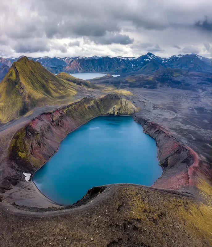 Aerial view of a vibrant blue crater lake, surrounded by colorful volcanic slopes under a cloudy sky.