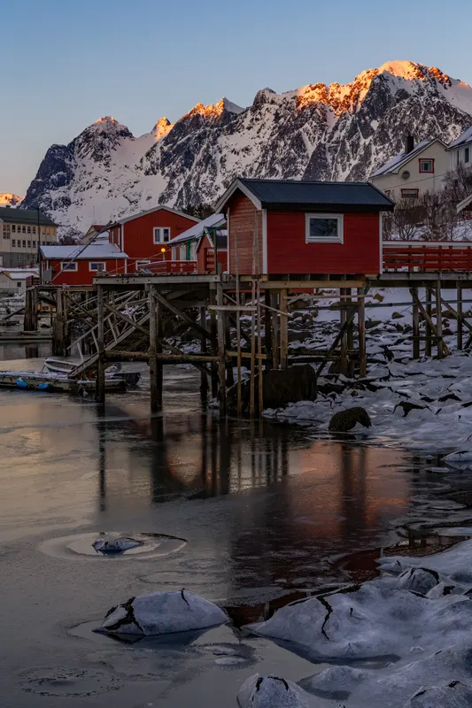 Rote Fischerhütten auf Stelzen am teilweise zugefrorenen Wasser, mit schneebedeckten Bergen im Hintergrund, deren Gipfel golden leuchten.
