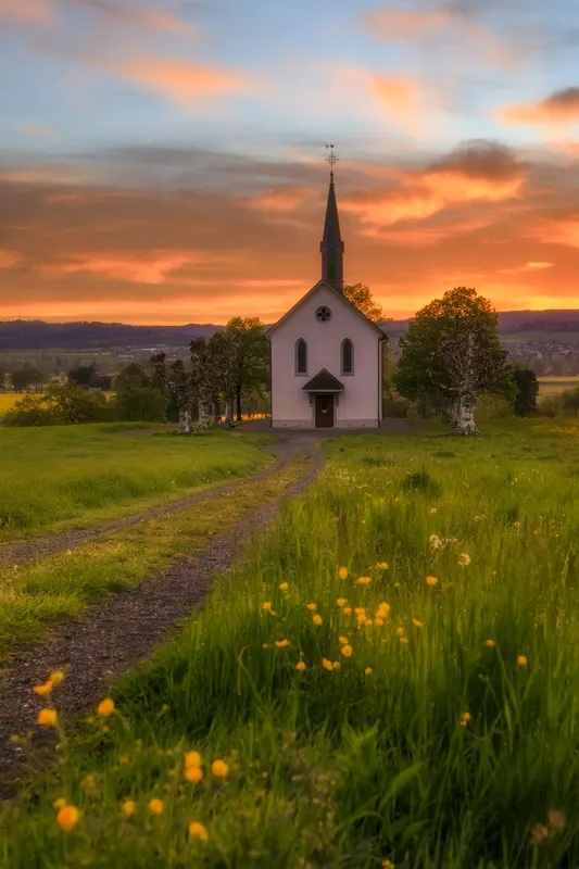 A white chapel with a pointed spire in a green meadow, illuminated by the golden light of a sunset with an orange sky.