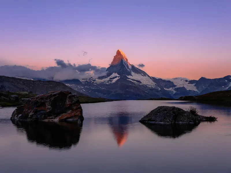 The Matterhorn at sunrise, its peak illuminated in gold, reflected in a mountain lake with rocks in the foreground.