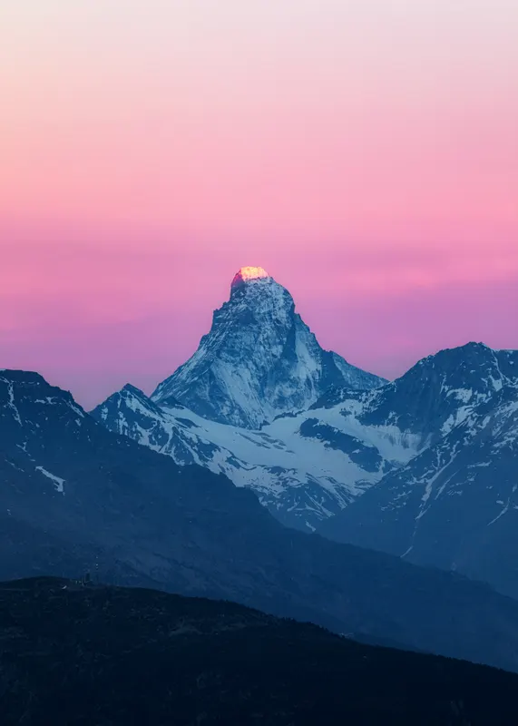 A snow-capped mountain with a golden glowing peak under a pink sky at sunrise.
