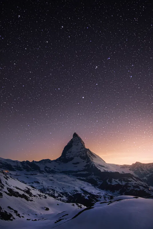 The Matterhorn at night, surrounded by snow-covered mountains under a clear starry sky with a bright horizon glow.