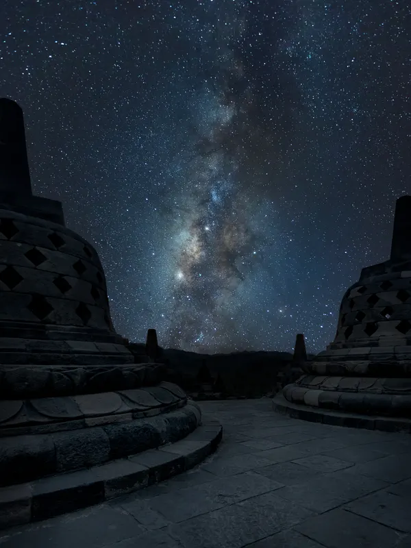 Nachtaufnahme der Milchstrasse über den dunklen, steinernen Stupas des Borobudur Tempels, die den Himmel einrahmen.