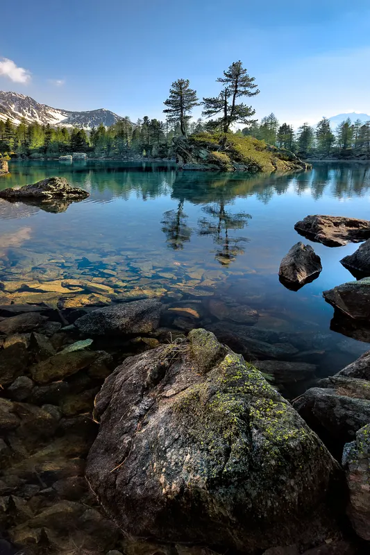 A clear mountain lake with visible rocks on the bottom, a small island with trees, and snow-capped mountains in the background.