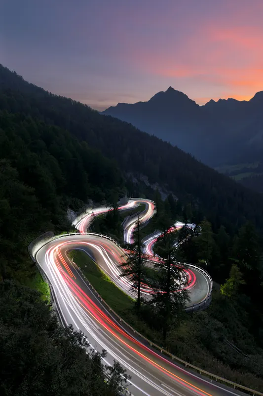 A long exposure image showing car light trails on a winding mountain road at dusk, surrounded by forests and mountains.
