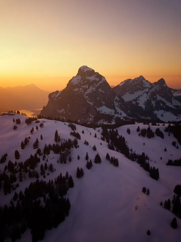 View over snow-covered hills with fir trees to the Mythen peaks at sunset, with Lake Lucerne in the background.