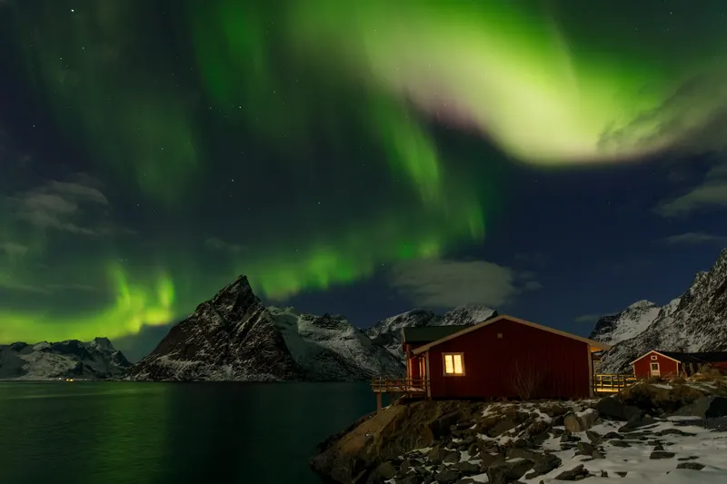 Green Northern Lights illuminate a fjord scene with snow-covered mountains and red fishing cabins, their windows glowing warmly.