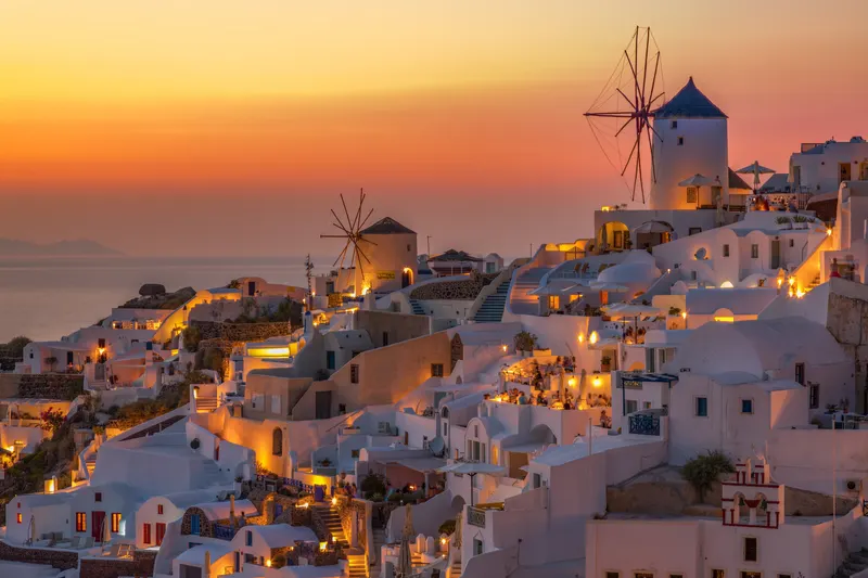 Abendansicht des Dorfes Oia auf Santorini mit weissen Häusern, Windmühlen und Lichtern, die unter einem orangefarbenen Himmel leuchten.