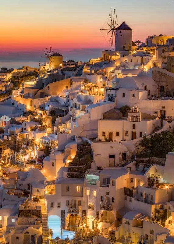 Eine Ansicht des Dorfes Oia auf Santorin bei Sonnenuntergang, mit weissen Häusern, die von Lichtern erhellt werden, und zwei Windmühlen vor einem farbigen Himmel.
