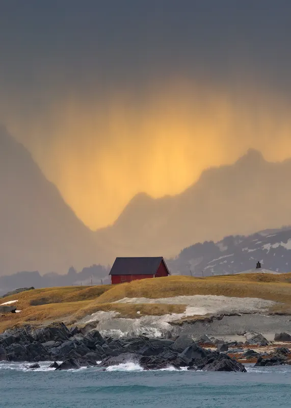 Eine rote Hütte auf einem grasbewachsenen Hügel an einer felsigen Küste, mit Bergen im Hintergrund und einem goldenen Himmel.