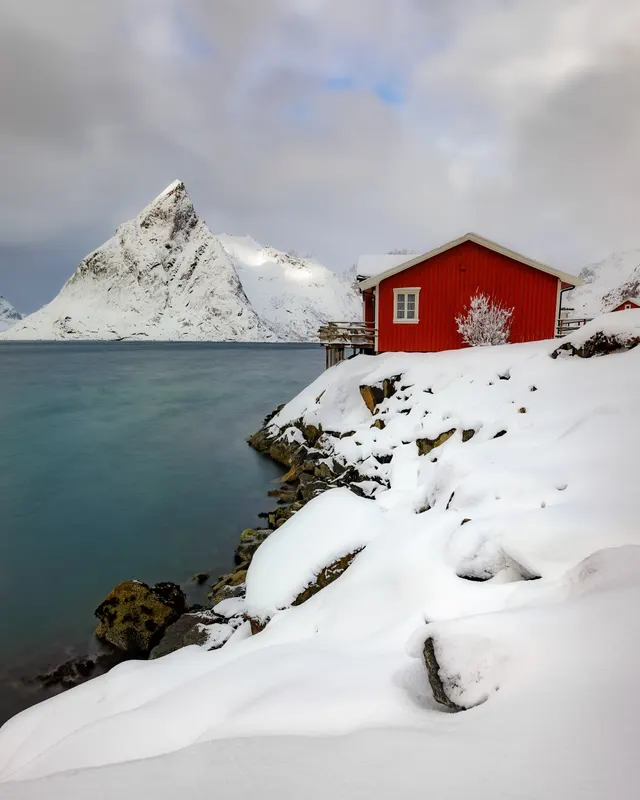 Eine rote Hütte am Ufer eines schneebedeckten Fjords mit einem spitzen, schneebedeckten Berg im Hintergrund unter einem bewölkten Himmel.