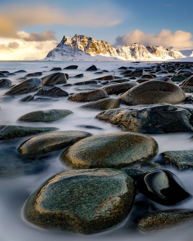 Winter coast in Norway: smooth rocks in milky water, sunlit snow-capped mountains in the background. Long exposure.