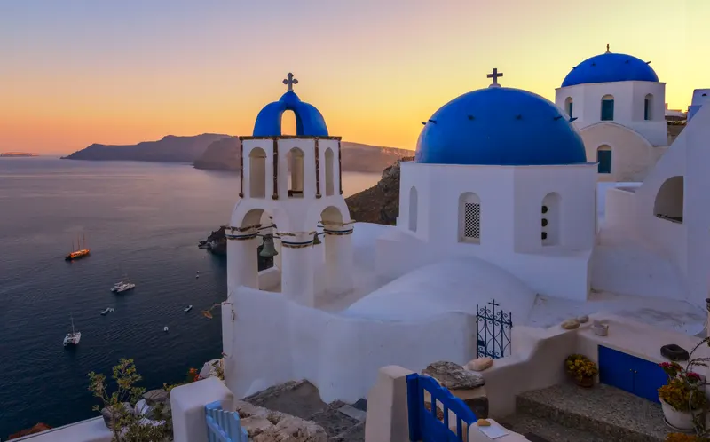 Blick auf weisse Kirchen mit blauen Kuppeln in Oia, Santorini, bei Sonnenuntergang über dem Meer und der Caldera.