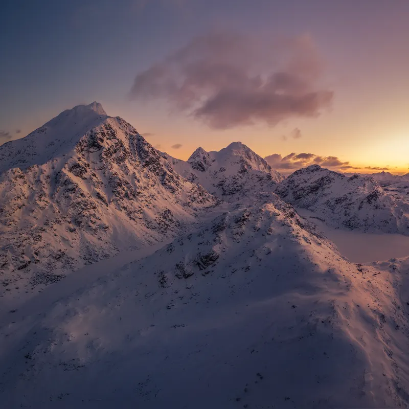 Panoramic image of a snow-covered mountain landscape at sunset, with warm colors on the horizon and cool shadows in the valleys.