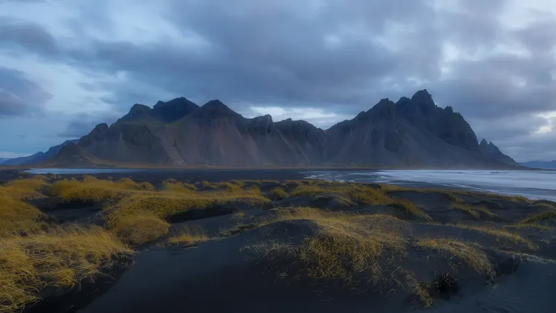A wide landscape with dark sand dunes and yellow grass in the foreground, a calm body of water, and a rugged mountain range under a cloudy sky.