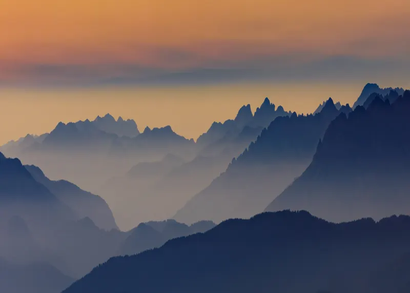 Panoramic image of layered mountain ranges at sunrise, fading from dark blue and grey into an orange sky.