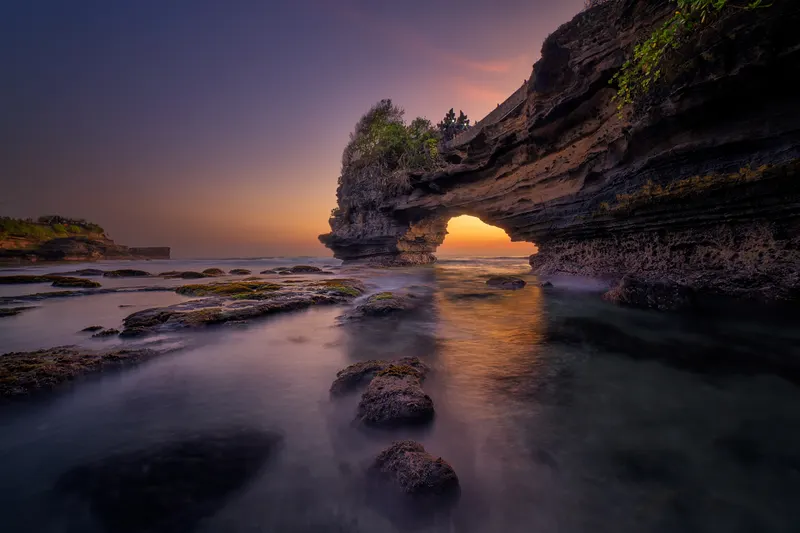 A natural rock arch by the sea at sunset, with calm water and a colorful sky.