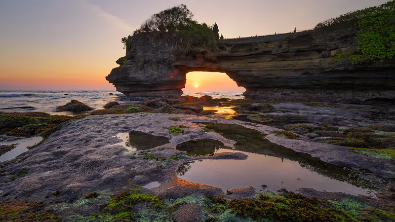 Sunset seen through a natural rock arch by the sea, with tidal pools in the foreground reflecting the warm light.
