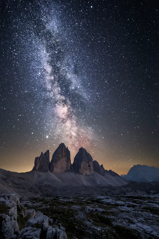 The Milky Way glows above the three distinctive peaks of Tre Cime di Lavaredo in a dark night landscape.