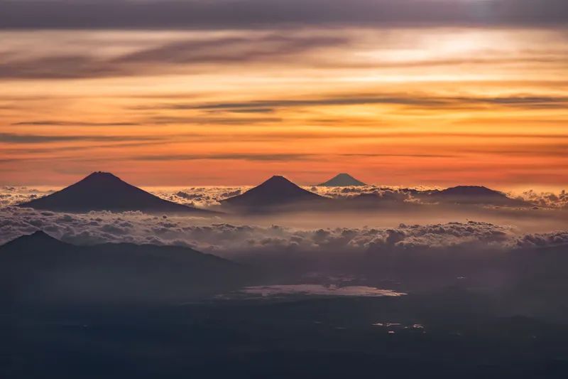Panoramablick auf mehrere Vulkane, die aus einem Wolkenmeer ragen, bei Sonnenaufgang mit orange-violettem Himmel.