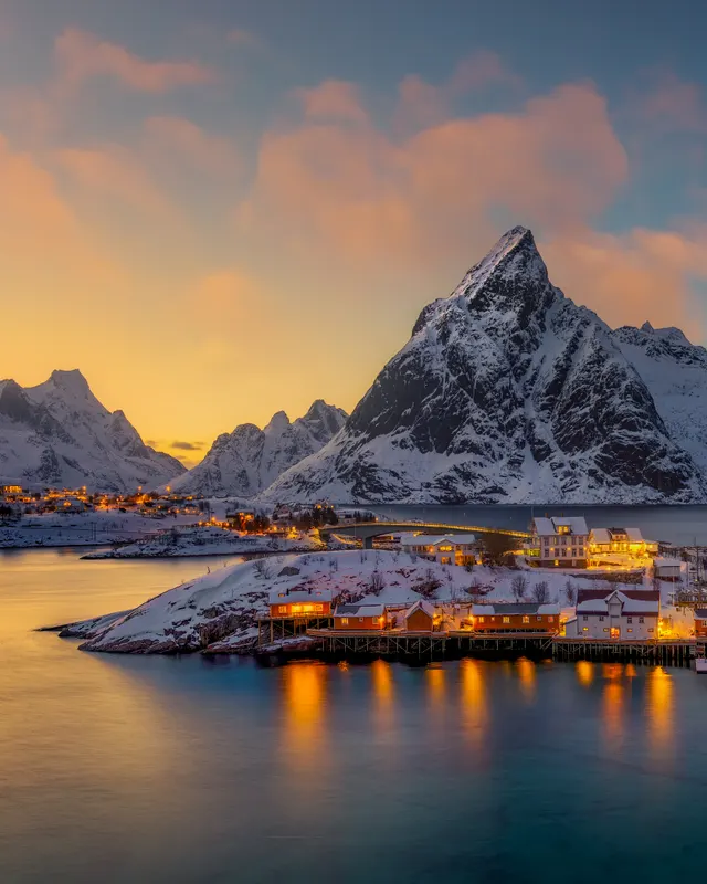 A winter view of the fishing village Reine in Norway at twilight, featuring illuminated houses reflecting in the water and snow-capped mountains in the background.