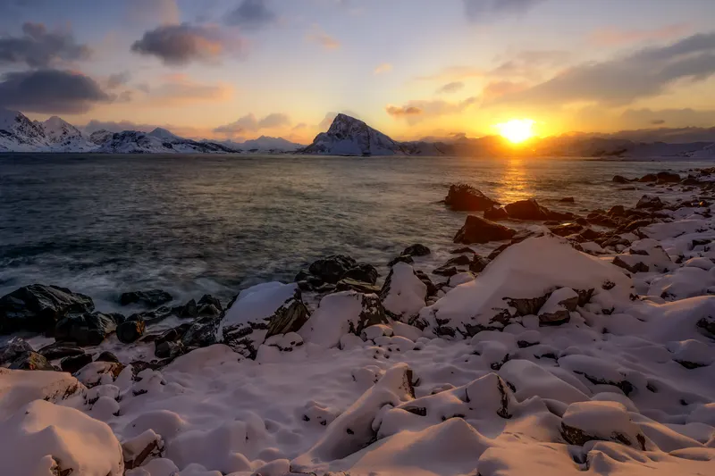 A winter sunset over a fjord in Norway, with snow-covered rocks in the foreground and mountains in the background.